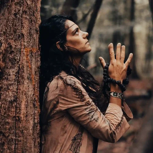 Femme brune adossée contre un arbre, les mains en prière avec un mala de rudra entre les mains